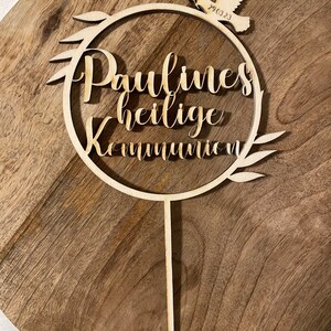 May include: A wooden cake topper with the words "Paulines heilige Kommunion" in script lettering. A dove with a sprig and the date "29.03.23" is at the top. The topper is on a wooden surface.