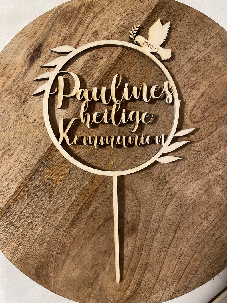 May include: A wooden cake topper with the words "Paulines heilige Kommunion" in script lettering. A dove with a sprig and the date "29.03.23" is at the top. The topper is on a wooden surface.