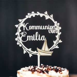 May include: A white wooden cake topper with the words "Kommunion von Emilia" in a circular floral design, and a small sailboat. The topper is placed on top of a cake with chocolate sprinkles.