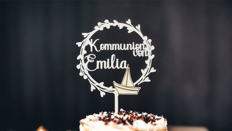 May include: A white wooden cake topper with the words "Kommunion von Emilia" in a circular floral design, and a small sailboat. The topper is placed on top of a cake with chocolate sprinkles.