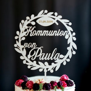 May include: A wooden cake topper with a wreath design, featuring the words "Kommunion von Paula" and the date 21.04.2024. The topper is placed on a white cake decorated with berries. The background is dark.