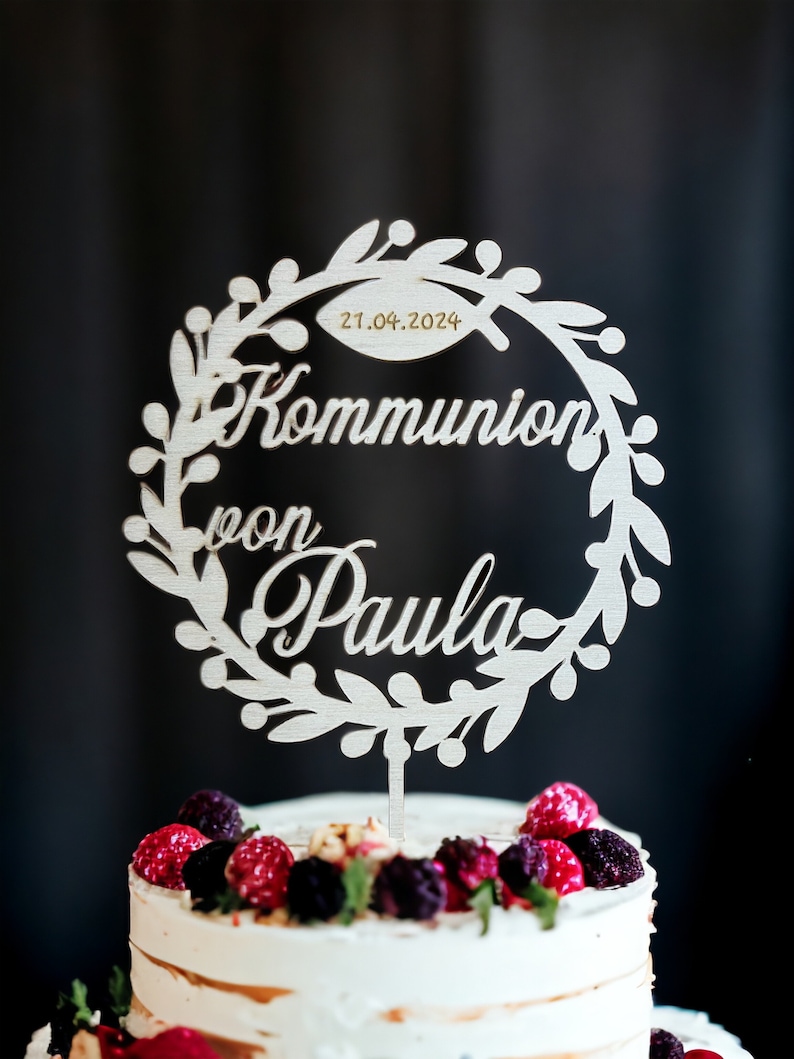 May include: A wooden cake topper with a wreath design, featuring the words "Kommunion von Paula" and the date 21.04.2024. The topper is placed on a white cake decorated with berries. The background is dark.