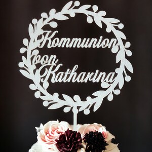 May include: A white cake topper with the words "Kommunion von Katharina" in script, surrounded by a floral wreath. The cake is topped with pink and burgundy flowers, set against a dark background.