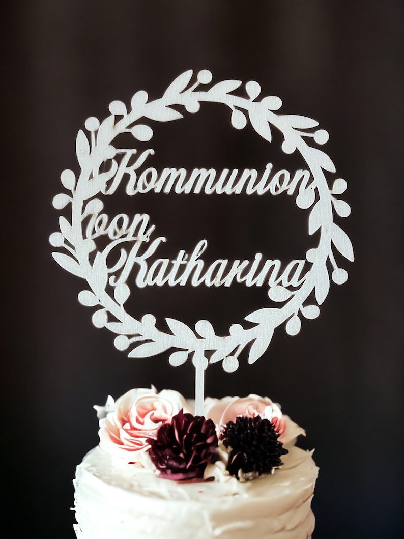 May include: A white cake topper with the words "Kommunion von Katharina" in script, surrounded by a floral wreath. The cake is topped with pink and burgundy flowers, set against a dark background.