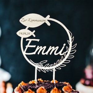 May include: A wooden cake topper with the name "Emmi" in cursive script, encircled by a wreath and two fish. The text "Zur Kommunion" and the date "14.04.24" are also included. The topper is placed on a cake.