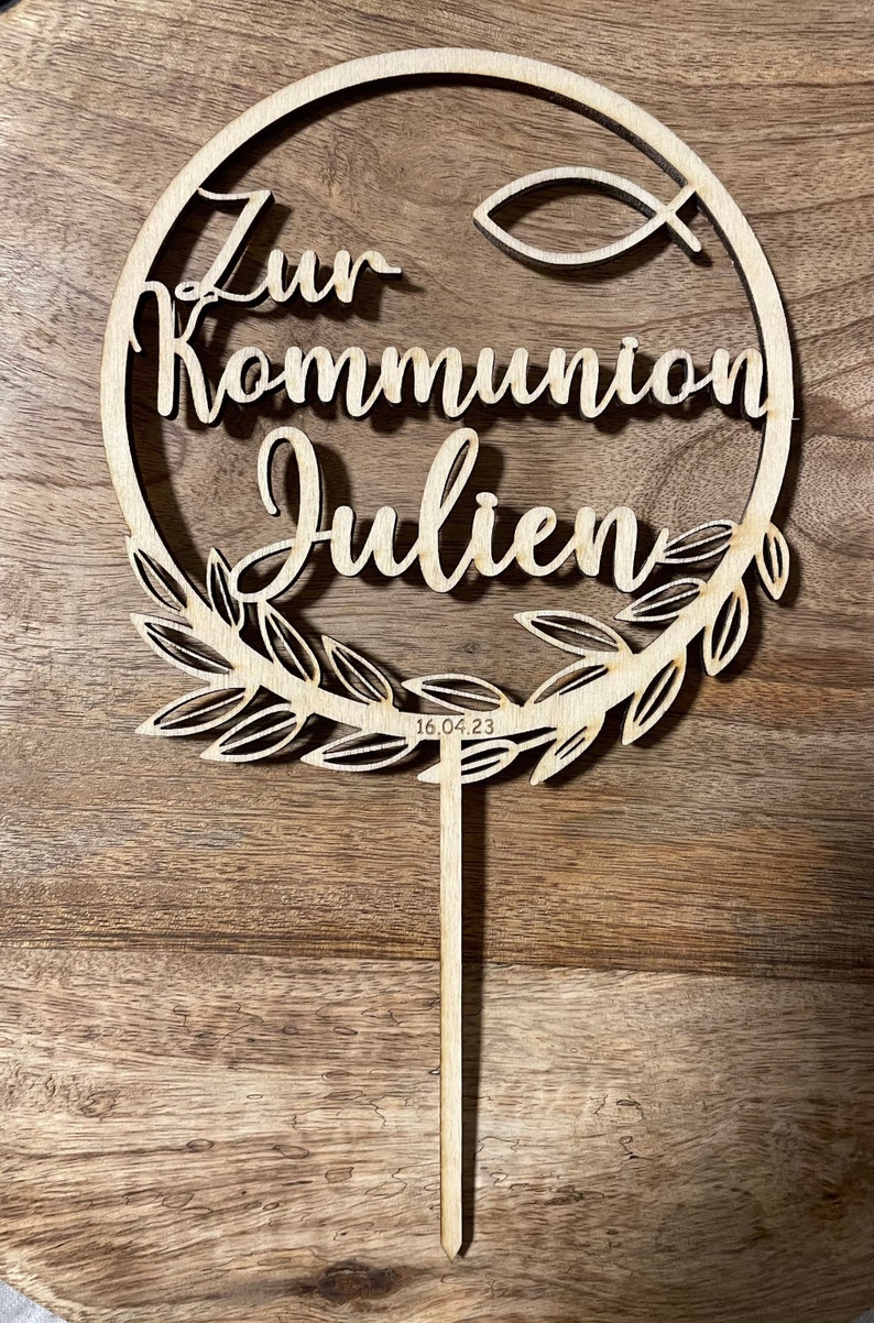 Kommunionscaketopper aus Holz mit einem Zweig am Rand und einem kleine Fisch im Design. Der Schriftzug "Zur Kommunion-Name" ist mittig.