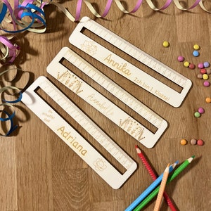 Könnte beinhalten: Drei Holzlineale mit eingravierten Namen und floralen oder Cartoon-Designs. Jedes Lineal hat eine Maßeinteilung bis 20 cm. Auf einem Lineal steht "Annika Ich bin 1. Klasse!" und auf einem anderen "Adriana Du schaffst das!"
