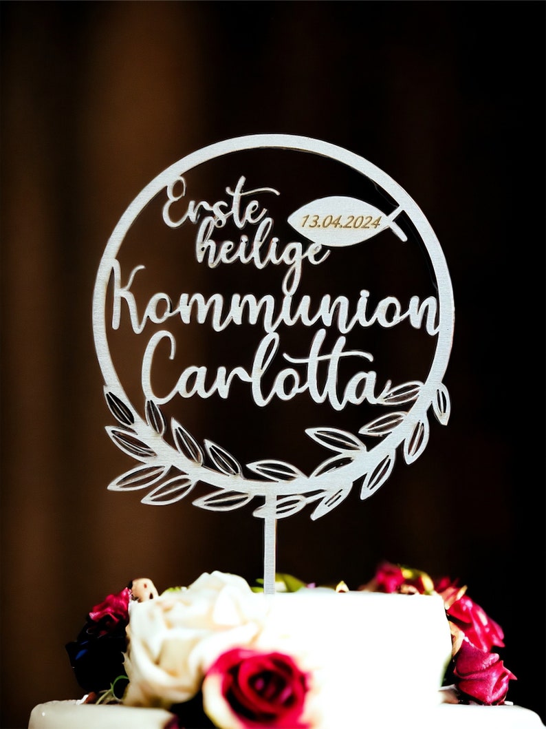 May include: A silver cake topper with the words "Erste heilige Kommunion Carlotta" in script lettering. The date "13.04.2024" is included. The topper is circular with a leaf design and is placed on a cake with red and white flowers.