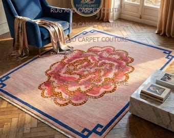 May include: A rectangular rug with a large floral design in shades of pink and gold. The rug has a light pink background with a dark blue border featuring geometric shapes. The rug is placed on a wooden floor.