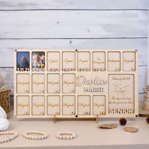 May include: A wooden photo frame with 12 slots for pictures. The frame is designed for a child's graduation, with the text "Devlin Marie" and "Class of 2039" printed on it. The frame also includes the text "The days are long, but the years are short" and "Senior".