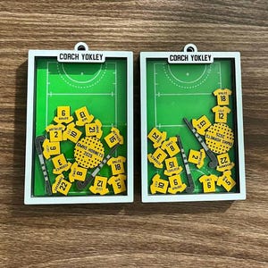 May include: Two rectangular ornaments with a white frame, featuring a green field hockey field design. Each ornament includes yellow jersey-shaped pieces with player numbers, hockey sticks, and the text "GMHS Hornets 2025" and "Coach Yokley".