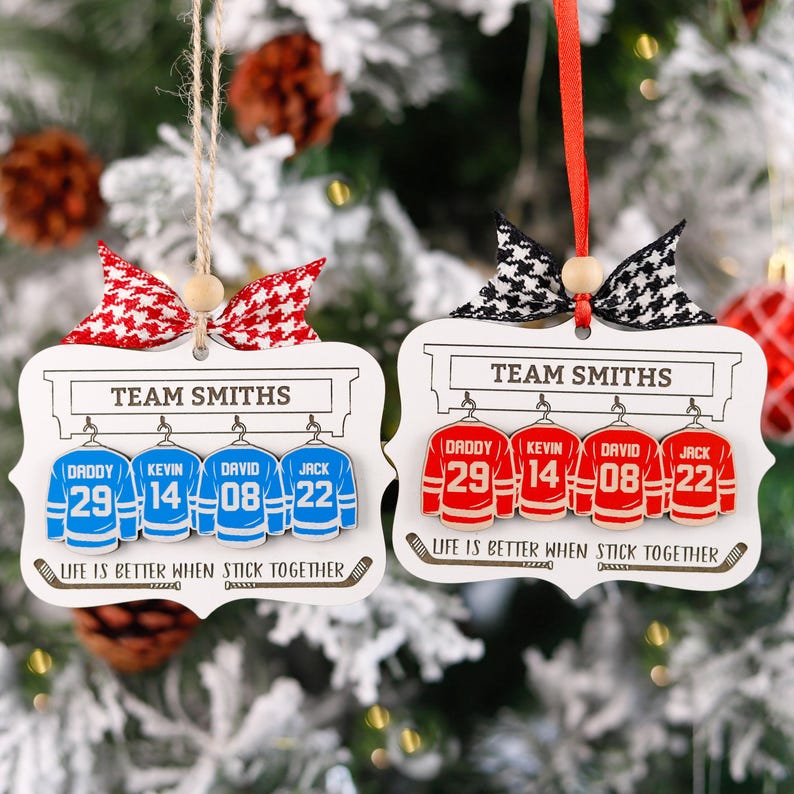 K&ouml;nnte beinhalten: Zwei wei&szlig;e Holzornamente in Form von Hockeytrikots mit dem Text "TEAM SMITHS" und "LIFE IS BETTER WHEN STICK TOGETHER". Die Trikots sind mit roten und blauen Hockeytrikots mit Nummern verziert. Die Ornamente h&auml;ngen an einem rot-wei&szlig;en Band.