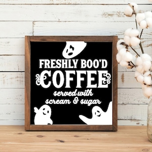 May include: A black wooden sign with white text that reads "Freshly Boo'd Coffee served with scream & sugar". There are three white ghost silhouettes on the sign.