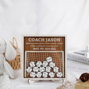 May include: A wooden sign with a soccer goal design and the text "COACH JASON" and "WHITE FOX 2024-2025". The sign has a quote about fearless players and coaches. There are 16 soccer balls with names and numbers on them.