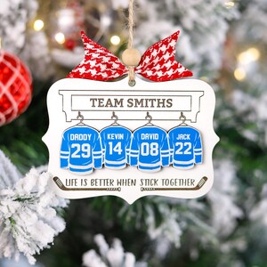 K&ouml;nnte beinhalten: Wei&szlig;er Weihnachtsbaumschmuck aus Holz mit Hockey-Motiv. Er zeigt vier Hockeytrikots mit den Namen "Daddy", "Kevin", "David" und "Jack" und ihren entsprechenden Nummern. Der Schmuck enth&auml;lt auch den Text "Team Smiths" und "Life is better when stick together".