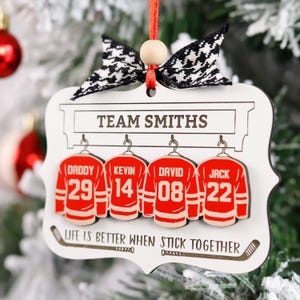 K&ouml;nnte beinhalten: Wei&szlig;er Weihnachtsbaumschmuck aus Holz mit Hockey-Motiv. Er zeigt vier rote Hockeytrikots mit den Namen "Daddy", "Kevin", "David" und "Jack" und ihren entsprechenden Nummern. Der Text "TEAM SMITHS" befindet sich oben und "LIFE IS BETTER WHEN STICK TOGETHER" unten.