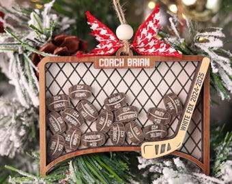 May include: Wooden hockey-themed Christmas ornament. Features a hockey net with a hockey stick, and small wooden barrels with names and the number 22. The ornament has the text "COACH BRIAN" and "WHITE FOX 2024-2025". Red and white patterned bow at the top.
