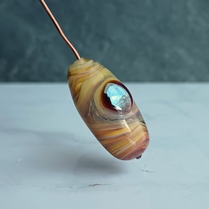 Può includere: Un perno di vetro con un motivo a spirale in tonalità di marrone, giallo e bianco. Il perno ha un piccolo cerchio blu iridescente al centro.