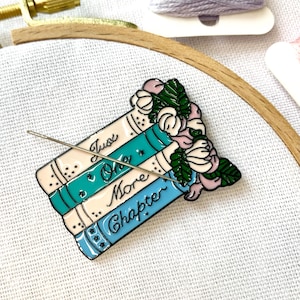Op de afbeelding: Een blauwe en witte emaille pin met een stapel boeken met de tekst "Just One More Chapter" en een bloemenontwerp. De pin is op een witte stoffen achtergrond.