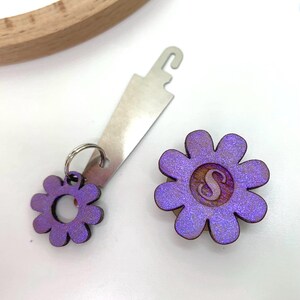 May include: Purple flower-shaped embroidery thread bobbin holder with a metal thread cutter. The bobbin holder has a hole in the center and the thread cutter has a hook on the end.