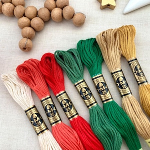 Puede incluir: Seis carretes de hilo de bordar DMC en varios colores, incluyendo blanco, rojo, naranja, verde y beige. Cada carrete tiene una etiqueta negra con el logotipo de DMC y el texto "DMC 25" y "8 m (8,7 yd)".