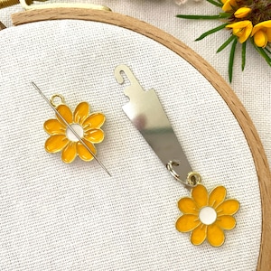 May include: A close-up of a needle threader with a yellow flower charm. The needle threader is made of metal and has a loop at the top. The flower charm is attached to the loop and has a white center.