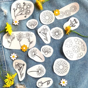 Könnte beinhalten: Eine Sammlung von Bügelbildern mit verschiedenen Gänseblümchen-Designs. Die Patches sind weiß mit schwarzen Umrissen und auf einem blauen Jeansstoff angeordnet.