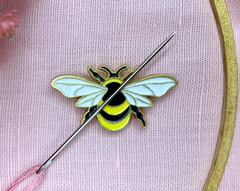 May include: A yellow and black bee pin with white wings is pinned to a pink fabric with a needle and thread going through it.