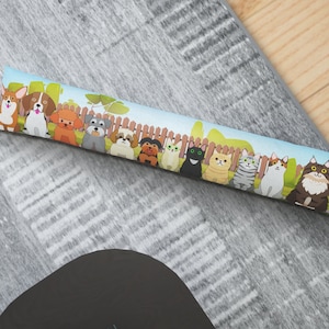 May include: A doorstop featuring a line of cartoon dogs and cats. The doorstop is made of fabric and has a light blue background with a white picket fence.