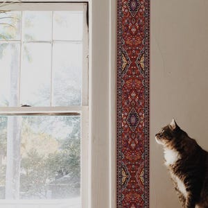 Può includere: Una finestra con vista su alberi e una casa in lontananza. La finestra è incorniciata da un bordo con motivo rosso e oro. Un gatto è seduto su una poltrona in pelle marrone, guardando fuori dalla finestra.