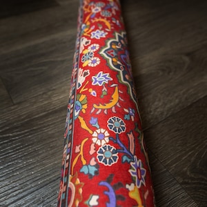 May include: A rolled-up red rug with a colorful floral pattern. The rug has a border with a repeating design.