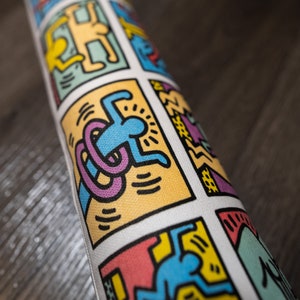 May include: A white yoga mat with a colorful repeating pattern of stick figures in various poses. The figures are outlined in black and filled with bright colors like yellow, blue, pink, and green.