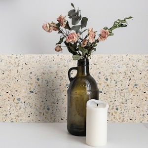 Puede incluir: Un jarrón de vidrio marrón con asa lleno de rosas rosadas secas y hojas de eucalipto. Una vela blanca se encuentra sobre una superficie blanca frente al jarrón. El fondo es un azulejo de terrazo beige.