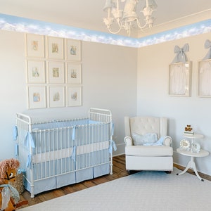 May include: A white crib with a blue and white bedding set, a white rocking chair with a blue and white pillow, and a white side table with a blue and white lamp. The room is decorated with a blue and white cloud border and a white chandelier with a rabbit figurine.