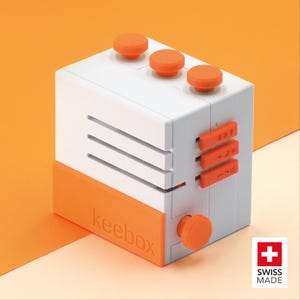 May include: A white and orange rectangular device labeled keebox with three circular buttons on top, three horizontal side sliders, and one circular knob on the side. The front panel features the embossed text keebox and a Swiss Made logo.