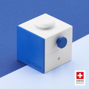 May include: A white and blue cubic device labeled keebox with a white circular button on the top surface and a blue circular knob on the side. The front blue panel features the text keebox embossed in a sans-serif font, and a small circular hole.
