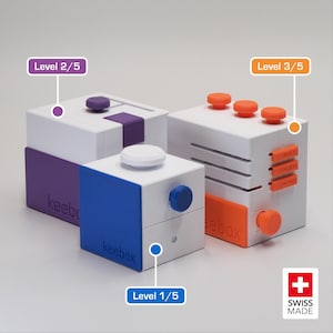 Pode incluir: Três cubos de manipulação de plástico branco rotulados como keebox com detalhes coloridos. Um cubo tem detalhes roxos e uma etiqueta Level 2/5, um tem detalhes azuis e Level 1/5, e um tem detalhes laranja e Level 3/5. Logotipo Swiss Made visível.