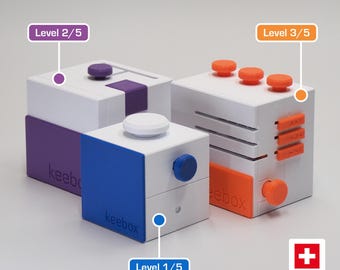 May include: Three white plastic fidget cubes labeled keebox with colored accents. One cube has purple details and a Level 2/5 tag, one has blue details and a Level 1/5 tag, and one has orange details and a Level 3/5 tag. A Swiss Made logo is visible.