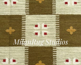 May include: Close-up of a woven rug with a repeating pattern. The design features squares of olive green and cream, with accents of red and gold. The text "Milan Rug Studios" is visible.