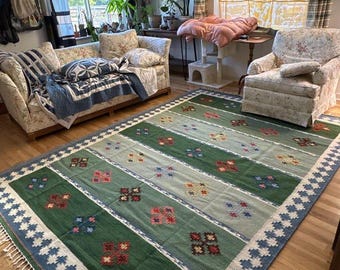 Puede incluir: Una alfombra rectangular con un patrón de rayas en tonos verdes, azules y crema. La alfombra presenta diseños florales y geométricos en rojo, rosa y azul. El borde tiene un patrón geométrico azul y blanco. La alfombra está colocada en un salón.