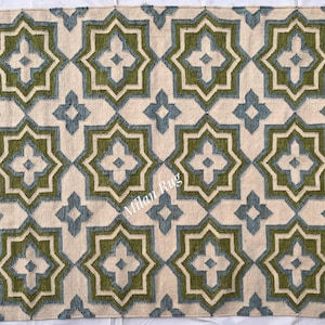 Può includere: Un tappeto beige con un motivo geometrico ripetuto nelle tonalità del verde e del blu. Il design presenta forme a stella a otto punte e motivi floreali più piccoli a quattro petali, creando un tessuto simmetrico e decorativo.