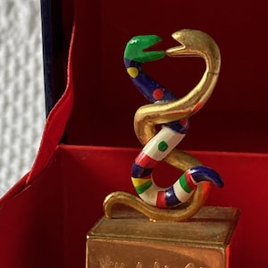 Puede incluir: Una escultura de serpiente dorada con detalles de esmalte de colores, sentada sobre una base dorada con el texto "Niki de Saint Phalle PRIMERA EDICI&Oacute;N".