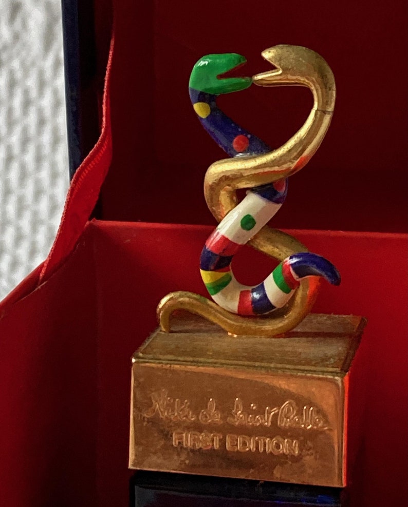 Puede incluir: Una escultura de serpiente dorada con detalles de esmalte de colores, sentada sobre una base dorada con el texto "Niki de Saint Phalle PRIMERA EDICI&Oacute;N".