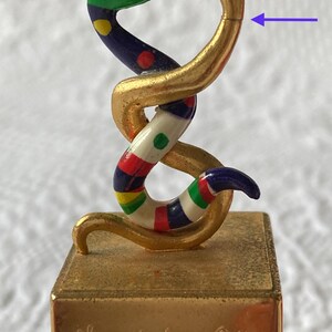 Puede incluir: Figurilla de serpiente de color dorado con una serpiente colorida y estampada envuelta alrededor. La base de la figurilla es dorada y tiene el texto "Nuit de Saint Phalle PRIMERA EDICI&Oacute;N" inscrito en ella.