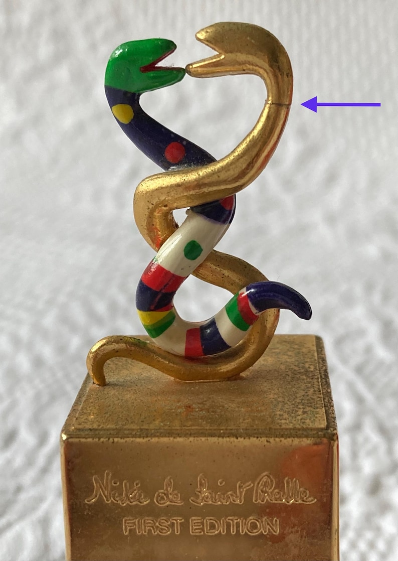 Puede incluir: Figurilla de serpiente de color dorado con una serpiente colorida y estampada envuelta alrededor. La base de la figurilla es dorada y tiene el texto "Nuit de Saint Phalle PRIMERA EDICI&Oacute;N" inscrito en ella.