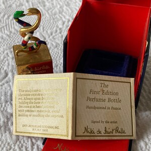 Puede incluir: Una escultura dorada de dos serpientes entrelazadas, con detalles de esmalte de colores, se asienta sobre una base cuadrada. La base est&aacute; inscrita con "Niki de Saint Phalle First Edition". La escultura est&aacute; en una caja roja con forro de terciopelo azul. La caja tambi&eacute;n incluye una tarjeta que dice "The First Edition Perfume Bottle Handpainted in France. Signed by the artist. Niki de Saint Phalle."