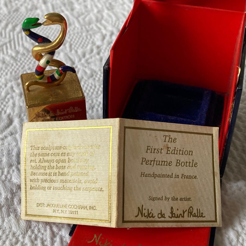 Puede incluir: Una escultura dorada de dos serpientes entrelazadas, con detalles de esmalte de colores, se asienta sobre una base cuadrada. La base est&aacute; inscrita con "Niki de Saint Phalle First Edition". La escultura est&aacute; en una caja roja con forro de terciopelo azul. La caja tambi&eacute;n incluye una tarjeta que dice "The First Edition Perfume Bottle Handpainted in France. Signed by the artist. Niki de Saint Phalle."