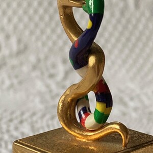 Puede incluir: Una escultura de serpiente de color dorado con un cuerpo colorido y estampado. La serpiente est&aacute; enroscada sobre s&iacute; misma y su cabeza est&aacute; levantada, con la boca abierta. La escultura est&aacute; sentada sobre una base cuadrada dorada.