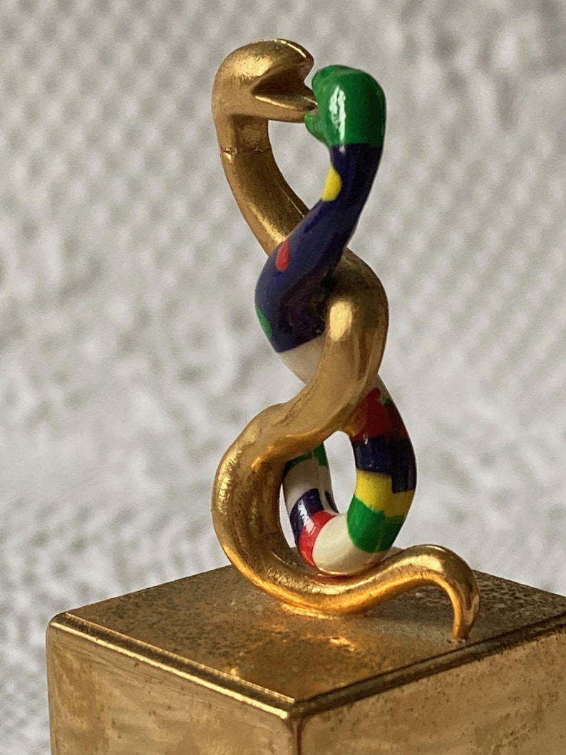 Puede incluir: Una escultura de serpiente de color dorado con un cuerpo colorido y estampado. La serpiente est&aacute; enroscada sobre s&iacute; misma y su cabeza est&aacute; levantada, con la boca abierta. La escultura est&aacute; sentada sobre una base cuadrada dorada.