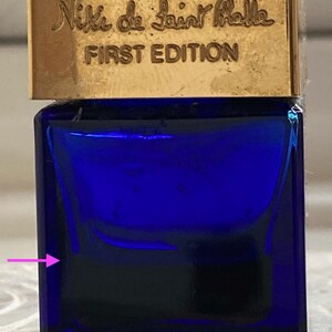 Puede incluir: Una peque&ntilde;a botella de vidrio azul con tapa dorada. La botella tiene el texto "Niki de Saint Phalle FIRST EDITION" en la parte superior.